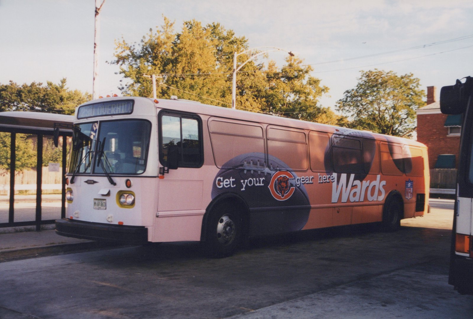 A white Pace bus with a Wards advertisement featuring a football and the Chicago Bears logo is parked at a terminal on a sunny day with autumn trees in the background.