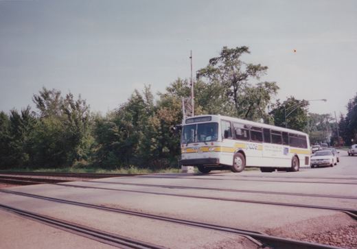 A white Pace bus with a yellow stripe, displaying route 209 Northwest, is driving over railroad tracks on a street, with green trees in the background.