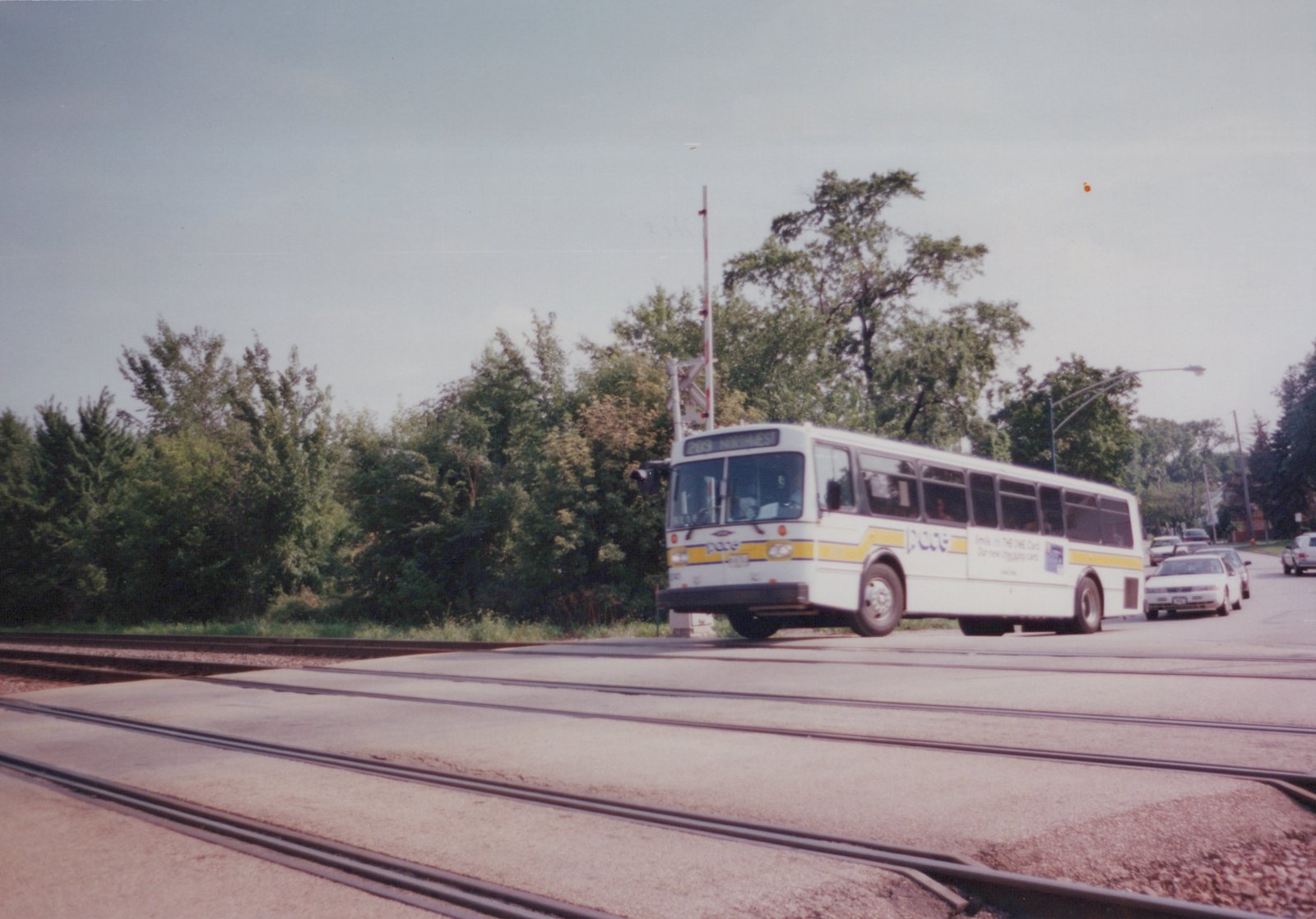 A white Pace bus with a yellow stripe, displaying route 209 Northwest, is driving over railroad tracks on a street, with green trees in the background.