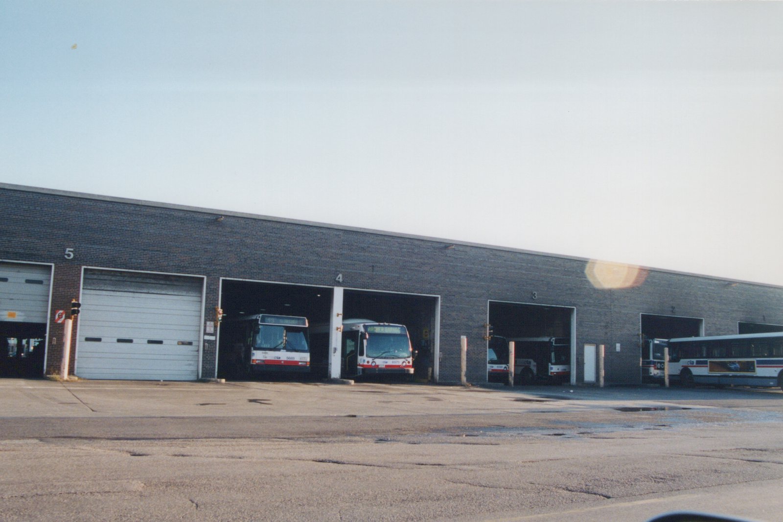 A row of white, red, and blue Chicago Transit Authority buses parked in front of a brick garage with numbered bays, with the sky overcast.