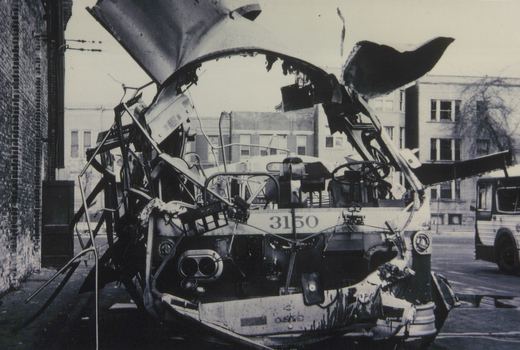 A black and white photo showing the front of a severely damaged, burned-out bus with the number 3150 on its front, parked on a city street with another bus in the background.
