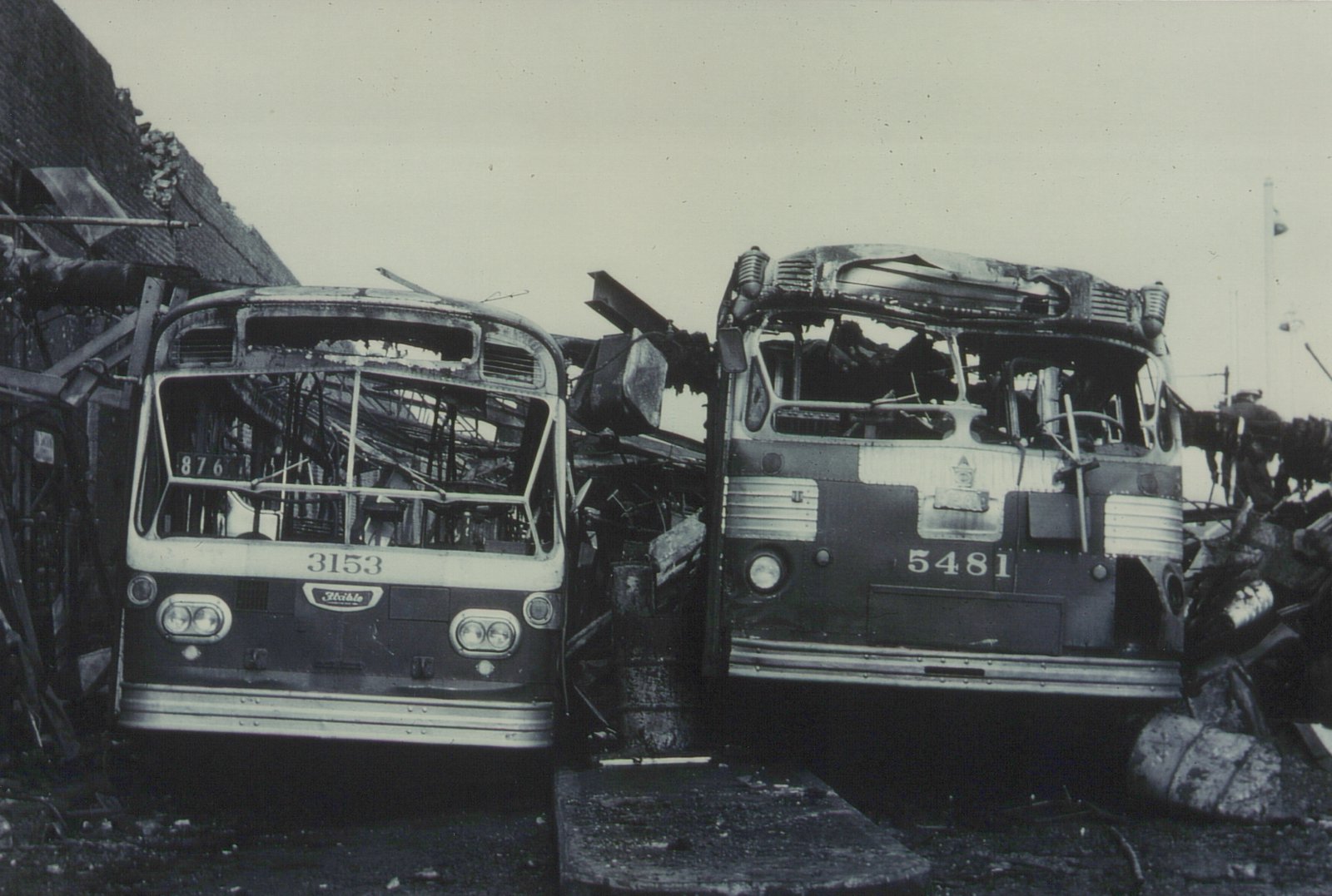 Two heavily damaged buses, numbered 3153 and 5481, stand side by side in a wrecked condition in a yard, with one showing the route number 876 on its destination sign.