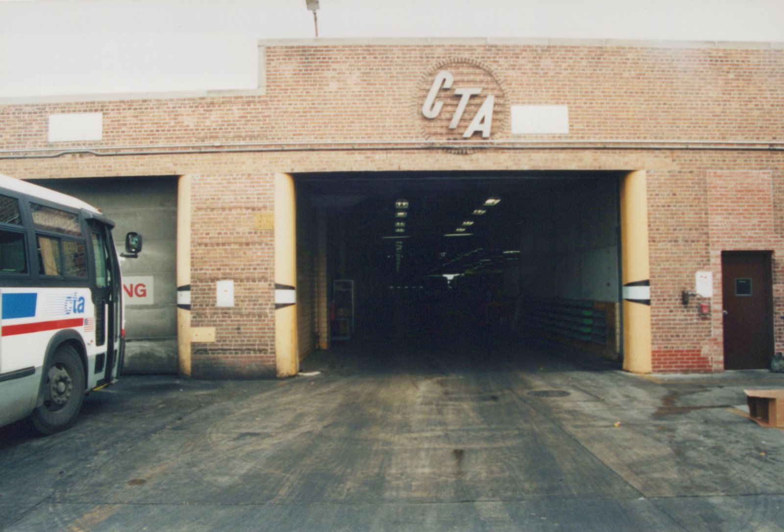 A white CTA bus with blue and red stripes is parked outside a brick garage with a large CTA logo above the entrance. The garage interior is dimly lit, and the ground is paved.