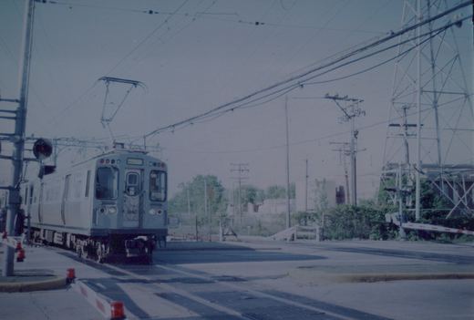 A silver and blue Chicago Transit Authority rapid transit car with the number 3450 on the front, stopped at a grade crossing with overhead wires and crossing gates.