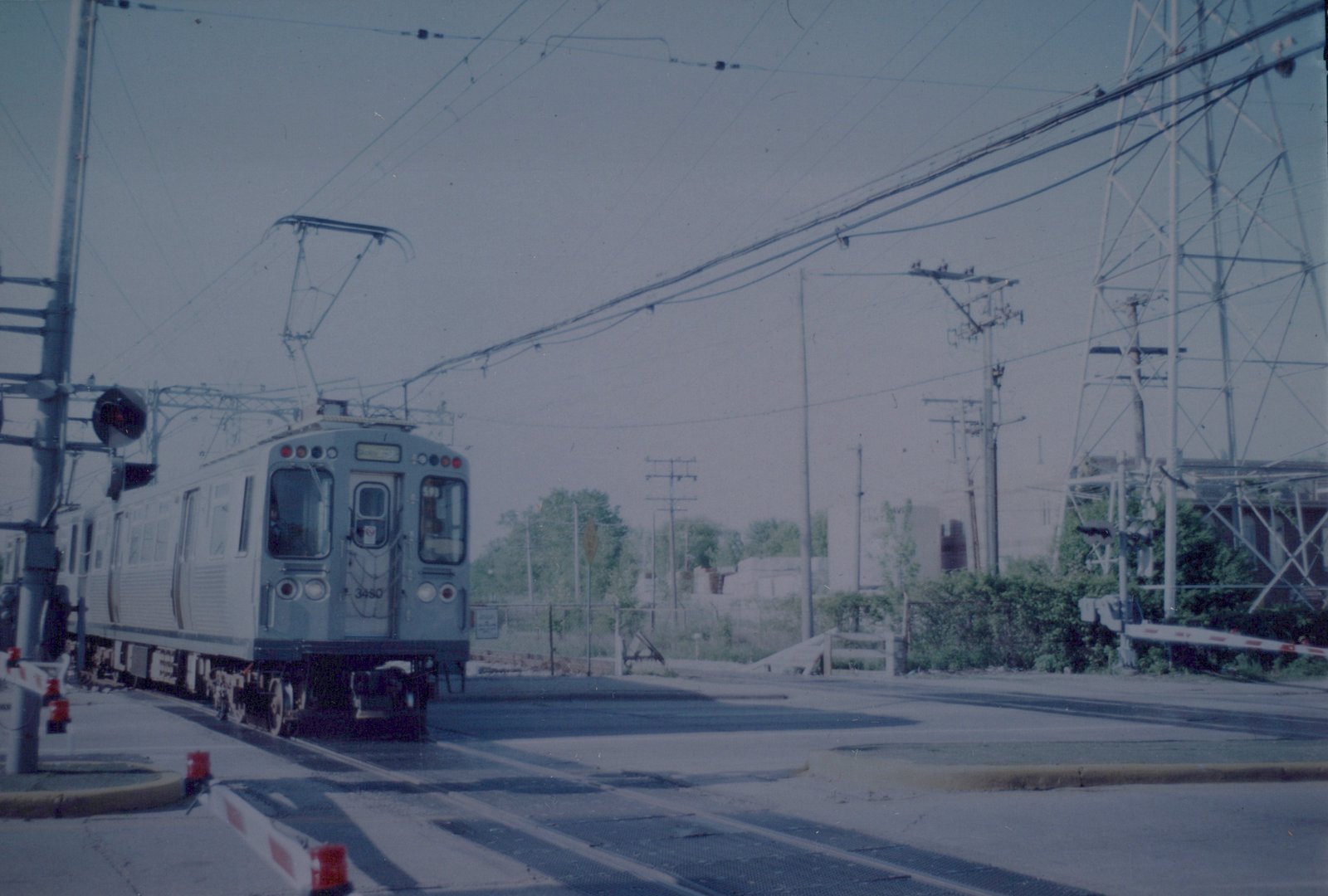 A silver and blue Chicago Transit Authority rapid transit car with the number 3450 on the front, stopped at a grade crossing with overhead wires and crossing gates.