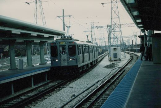 A silver CTA rapid transit train, number 594, with a 'Skokie' sign on the front, is stopped at a station platform. Three people wait on the platform under a shelter. The scene is overcast, with overhead electric wires visible.