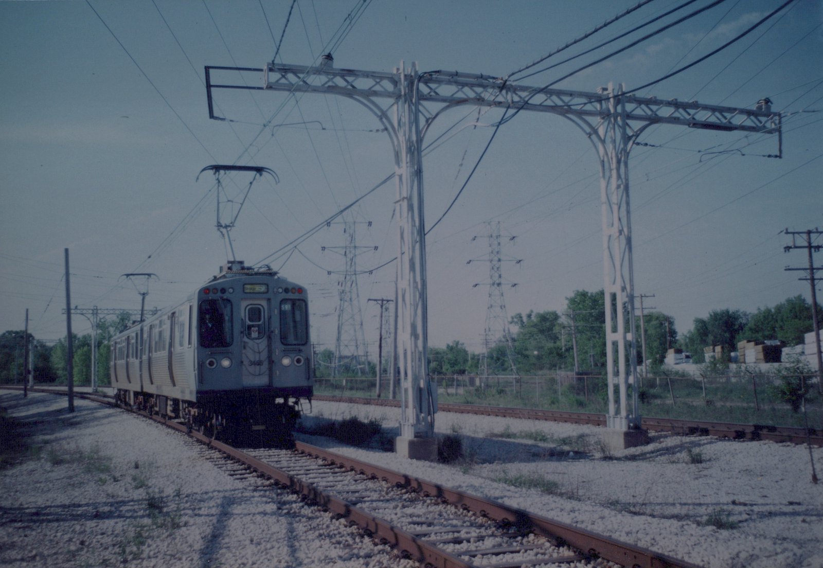 A silver rapid transit car with the number 3455 and 'SKOKIE' on its destination sign moves along tracks under overhead wires, with utility poles and green trees in the background.