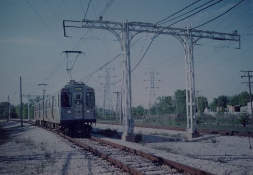 A silver rapid transit car with the number 3455 and 'SKOKIE' on its destination sign moves along tracks under overhead wires, with utility poles and green trees in the background.