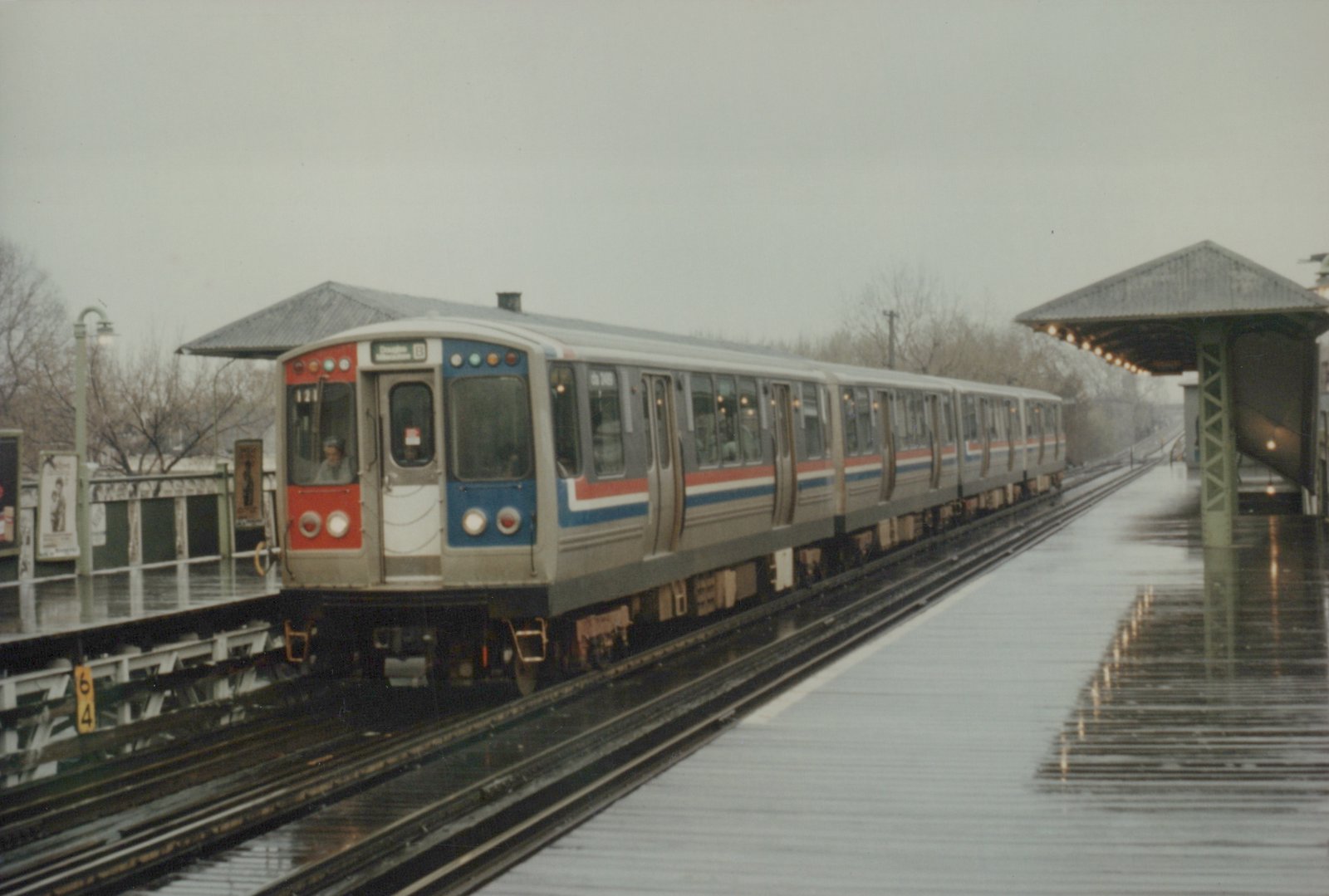 A silver rapid transit train with red and blue stripes, numbered 121, is stopped at an elevated station platform on a wet, overcast day.