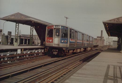 A Chicago Transit Authority 2000-series rapid transit car with gray body and red and blue stripes is stopped at an elevated station with wooden platforms and metal canopies on an overcast day.