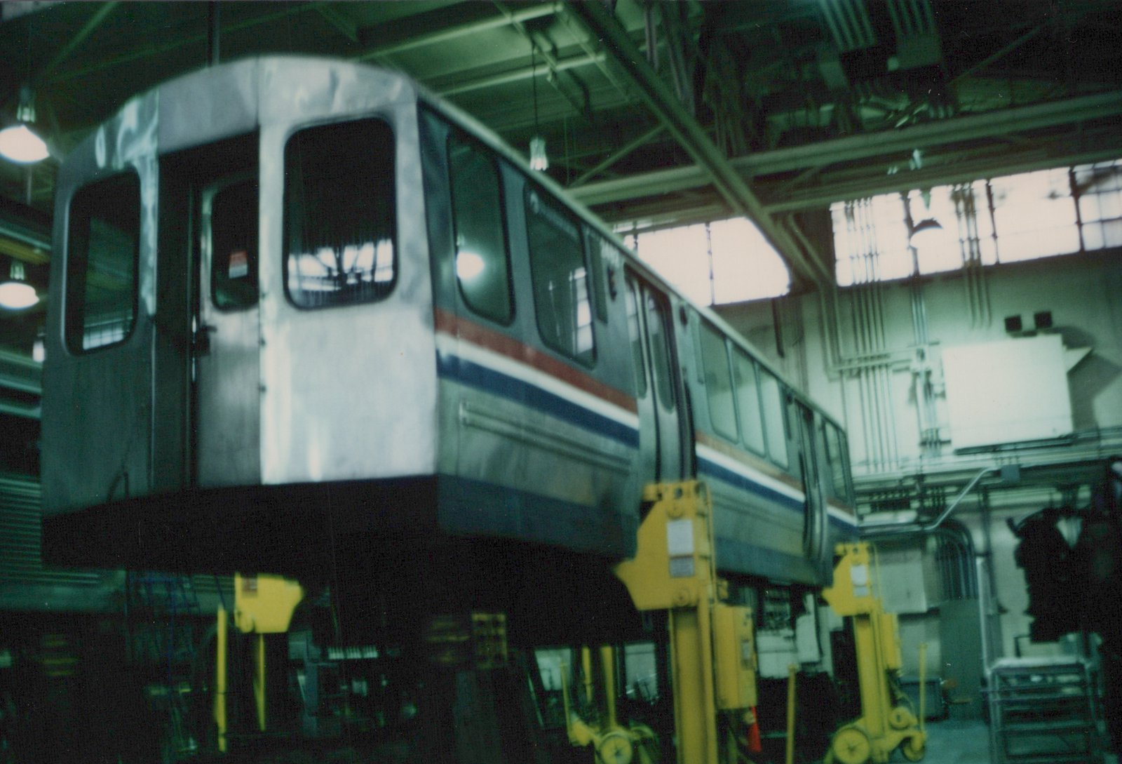 A silver CTA 2000-series subway car with red, white, and blue stripes is lifted on yellow hydraulic jacks inside a workshop with high windows and exposed ceiling beams.
