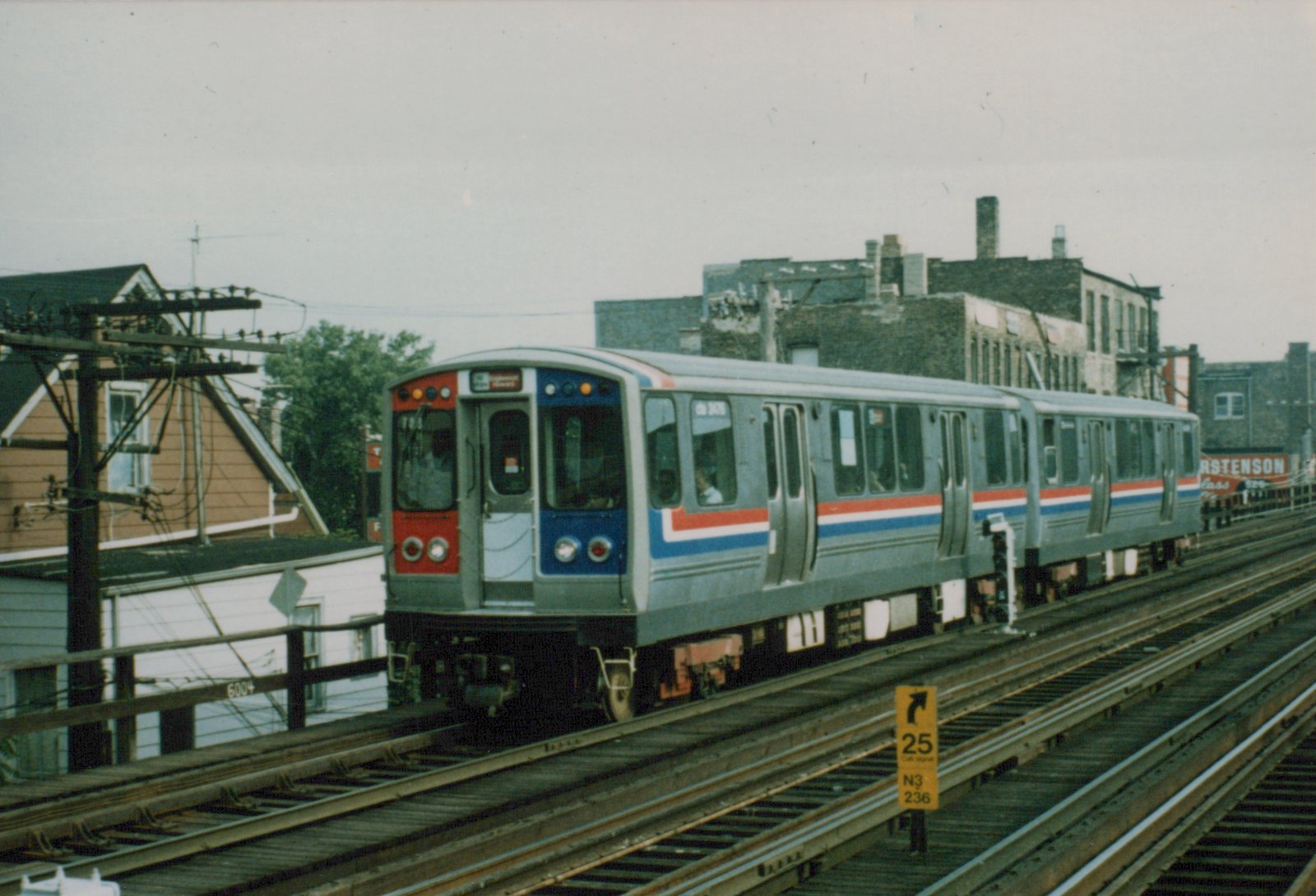 A red, white, and blue CTA rapid transit train on elevated tracks with buildings and a utility pole in the background.