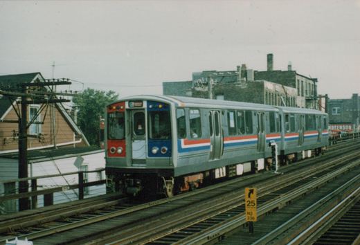 A red, white, and blue CTA rapid transit train on elevated tracks with buildings and a utility pole in the background.