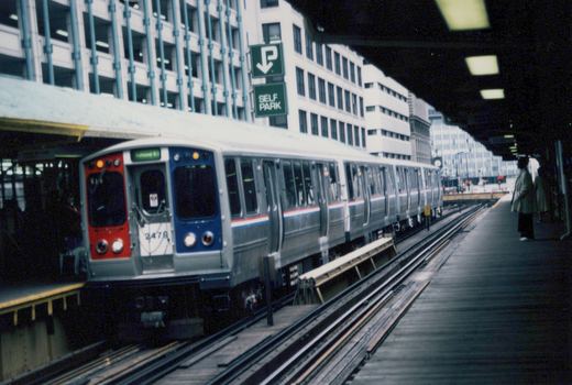 A silver, red, and blue Chicago 'L' train at an elevated station platform, with passengers waiting and a green 'SELF PARK' sign overhead.