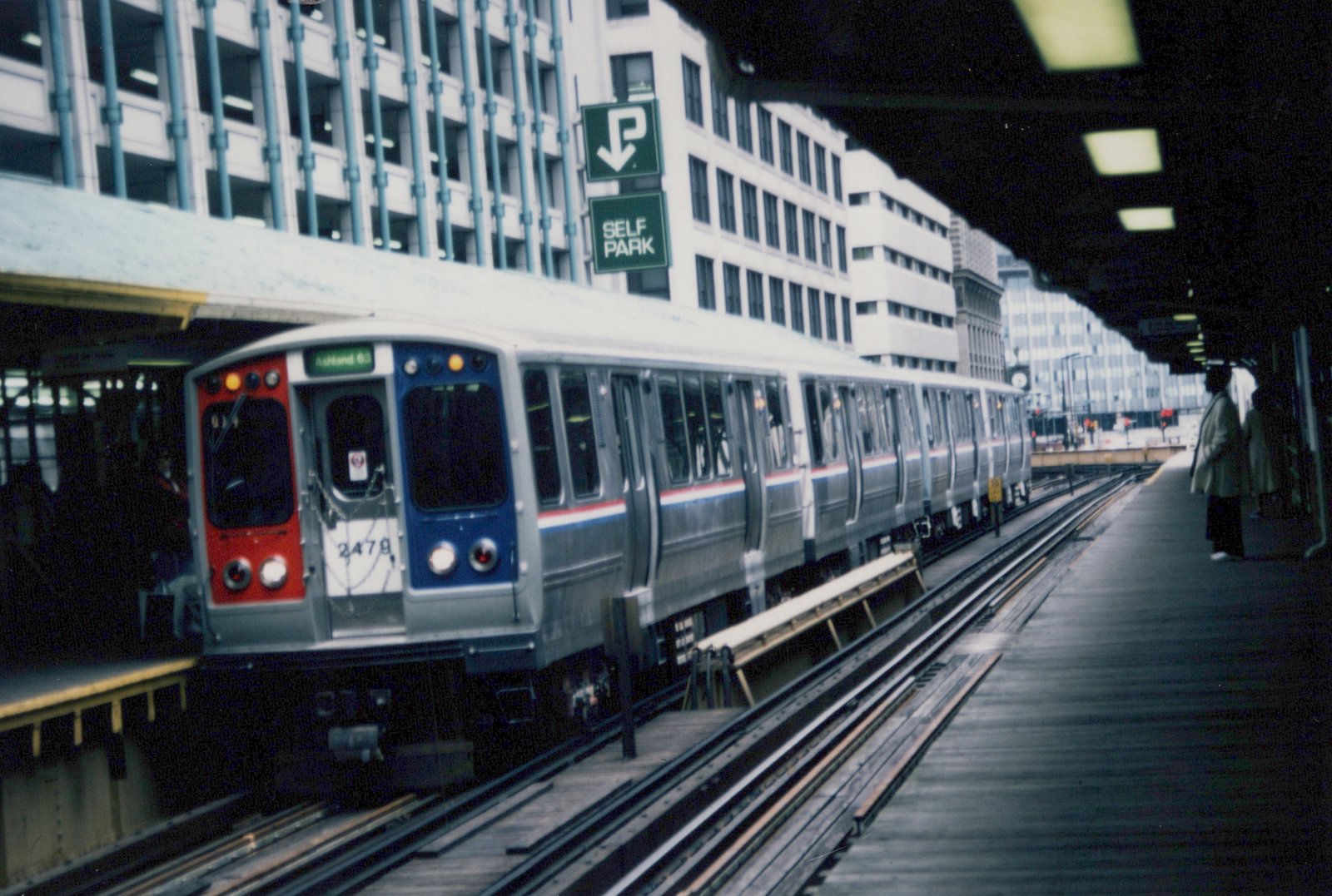 A silver, red, and blue Chicago 'L' train at an elevated station platform, with passengers waiting and a green 'SELF PARK' sign overhead.
