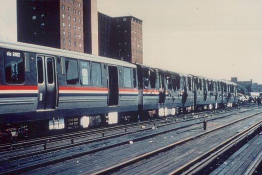 A long line of blue and red Chicago 'L' cars on an elevated track, with the lead car showing the number 2482 and 'cta' on its front window, against a backdrop of tall buildings.