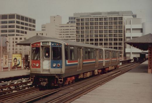 An elevated train car with red, white and blue stripes, labeled CTA 2460, stopped at a station platform in an urban setting with office buildings in the background.