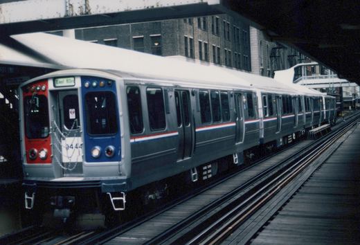 A silver Chicago Transit Authority rapid transit train with red and blue stripes, numbered 2444, sits on an elevated track under a steel bridge with a brick building in the background.