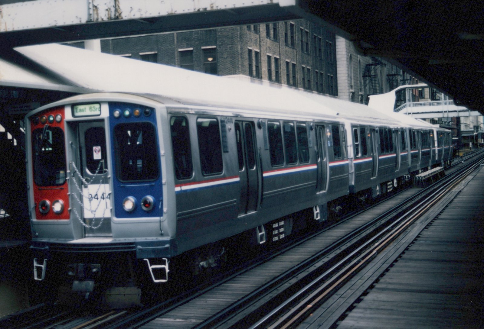 A silver Chicago Transit Authority rapid transit train with red and blue stripes, numbered 2444, sits on an elevated track under a steel bridge with a brick building in the background.