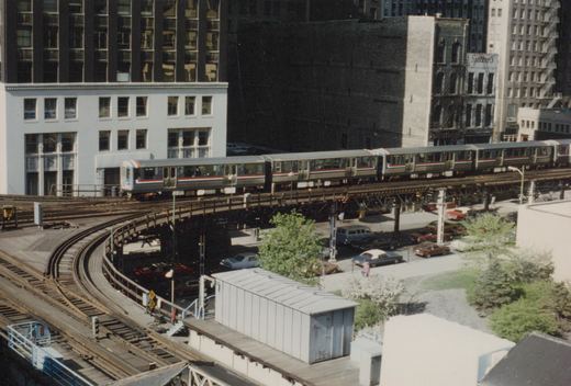 An elevated train with a silver body and red, white, and blue stripe travels on a curved track above a street with parked cars and buildings in the background, including one with a 'Dixon' sign and another with a 'Spitzer's' sign.