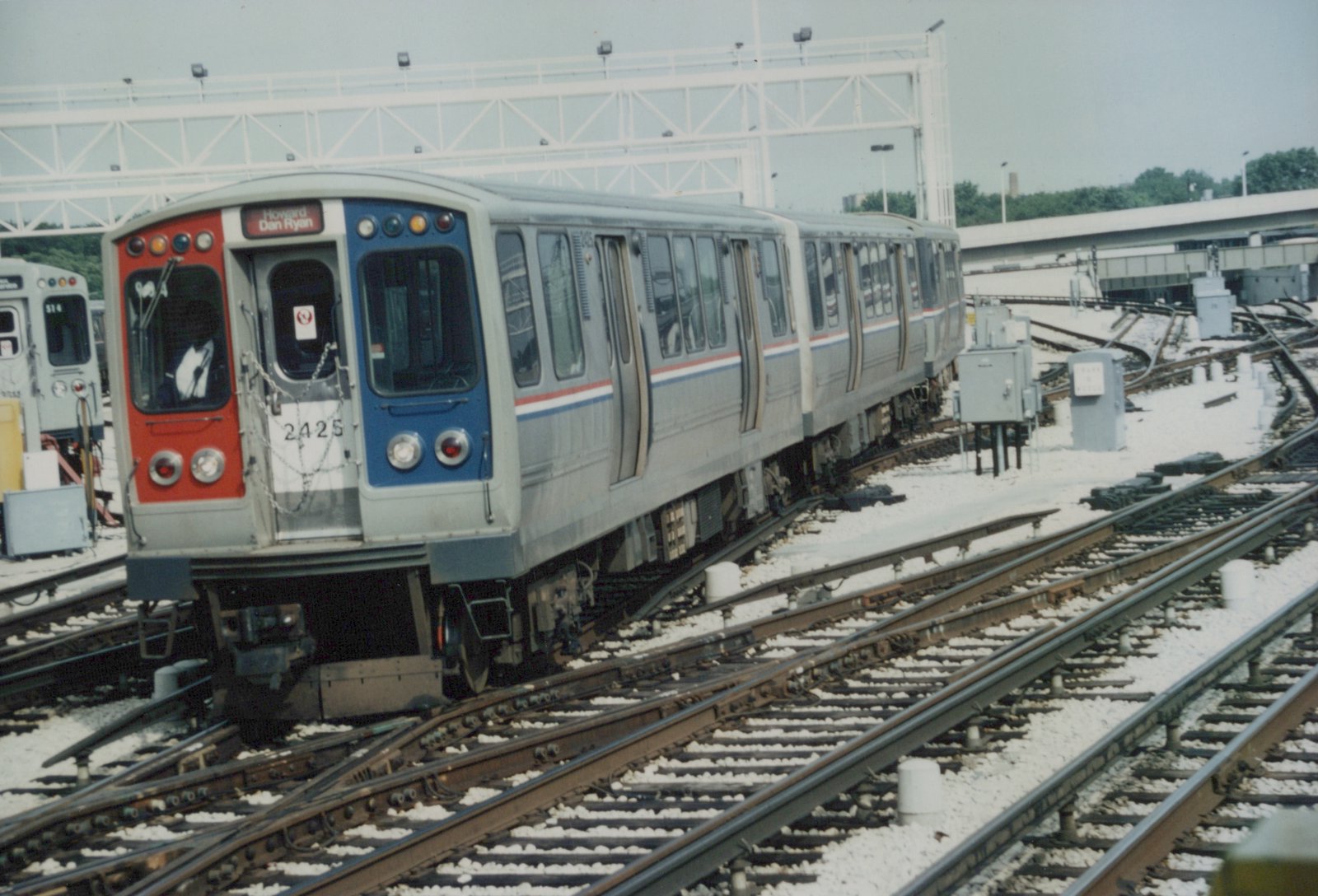 A silver Chicago Transit Authority rapid transit car with red, white, and blue stripes, number 2425, is positioned on tracks in a yard, with another car, number 514, visible in the background.