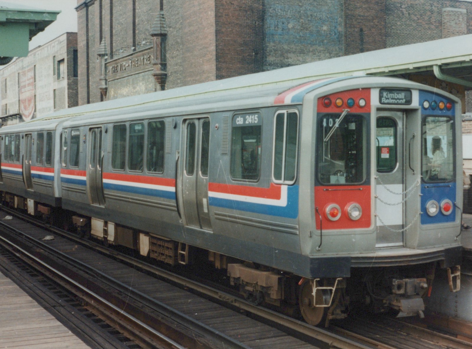 A silver CTA rapid transit car with red and blue stripes, numbered 2415, is stopped at an elevated station platform with a 'Kimball Belmont' sign on the front, and a brick building labeled 'THE VIC-THEATRE' in the background.