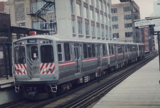 A silver Chicago 'L' train with red stripes, numbered 2406, is stopped at an elevated station platform under overcast skies. The front sign reads 'Evanston Express'.