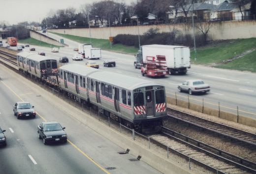 An elevated view of a silver and red Chicago 'L' train with the number 2404 on its front, running parallel to a highway with cars and trucks in the lanes beside it.