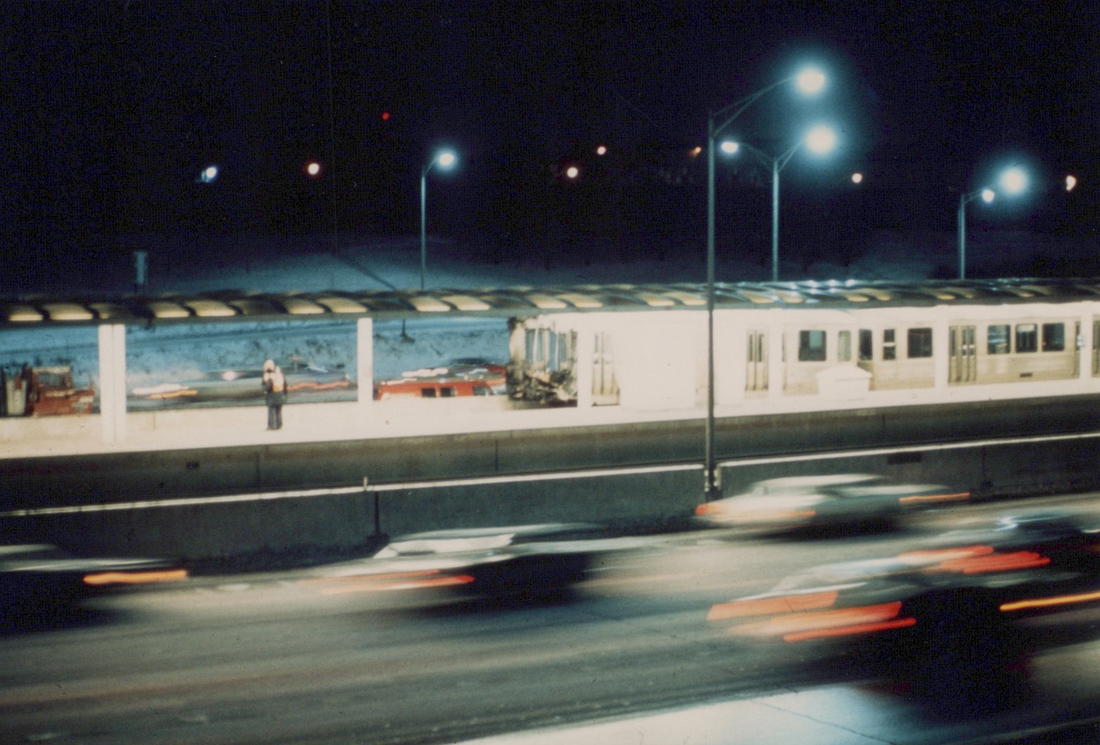 A night scene of a white rapid transit train on an elevated track, with a station platform and moving cars visible below.