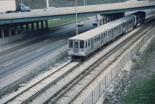 A silver CTA rapid transit train with the number 2260 travels on tracks next to a multi-lane highway, with cars and a truck on an overpass above.