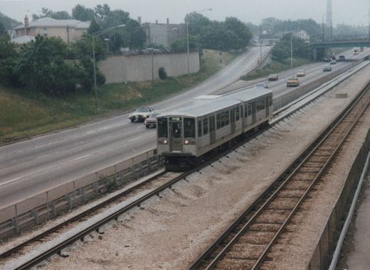 A silver CTA train with the number 2260 and destination 'O'HARE' travels on tracks beside a multi-lane highway with cars, viewed from an overpass on a cloudy day.