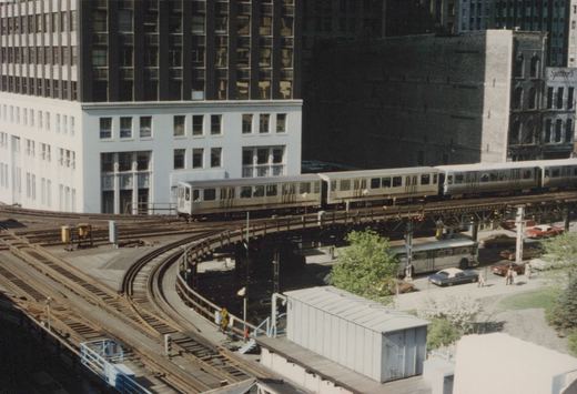 A silver CTA train with blue stripes travels on an elevated track, curving through a cityscape of tall buildings, with street-level traffic and pedestrians below.