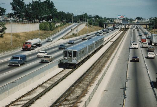 An elevated view of a silver CTA train running in the highway median next to cars from the 1980s, with a clear blue sky above.