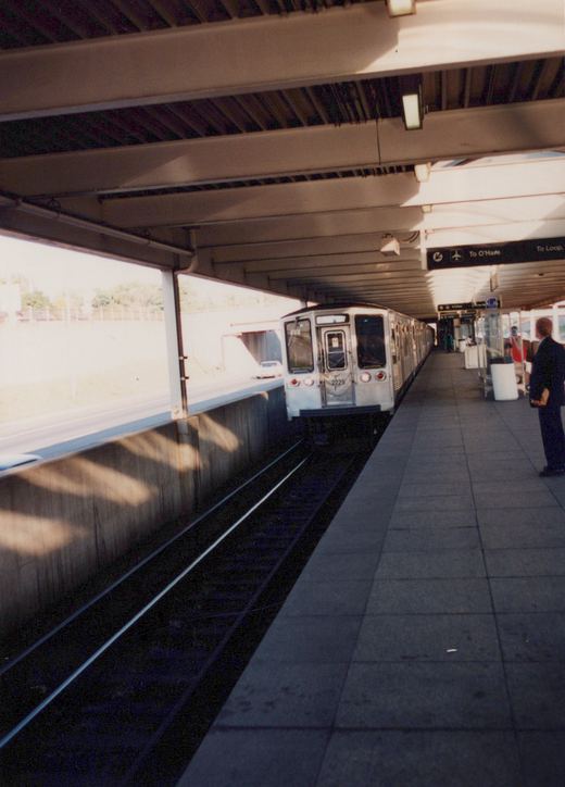 A silver CTA rapid transit train with the number 2225 on its front is stopped at a station platform under a covered structure; signs above the platform indicate directions to O'Hare and the Loop.