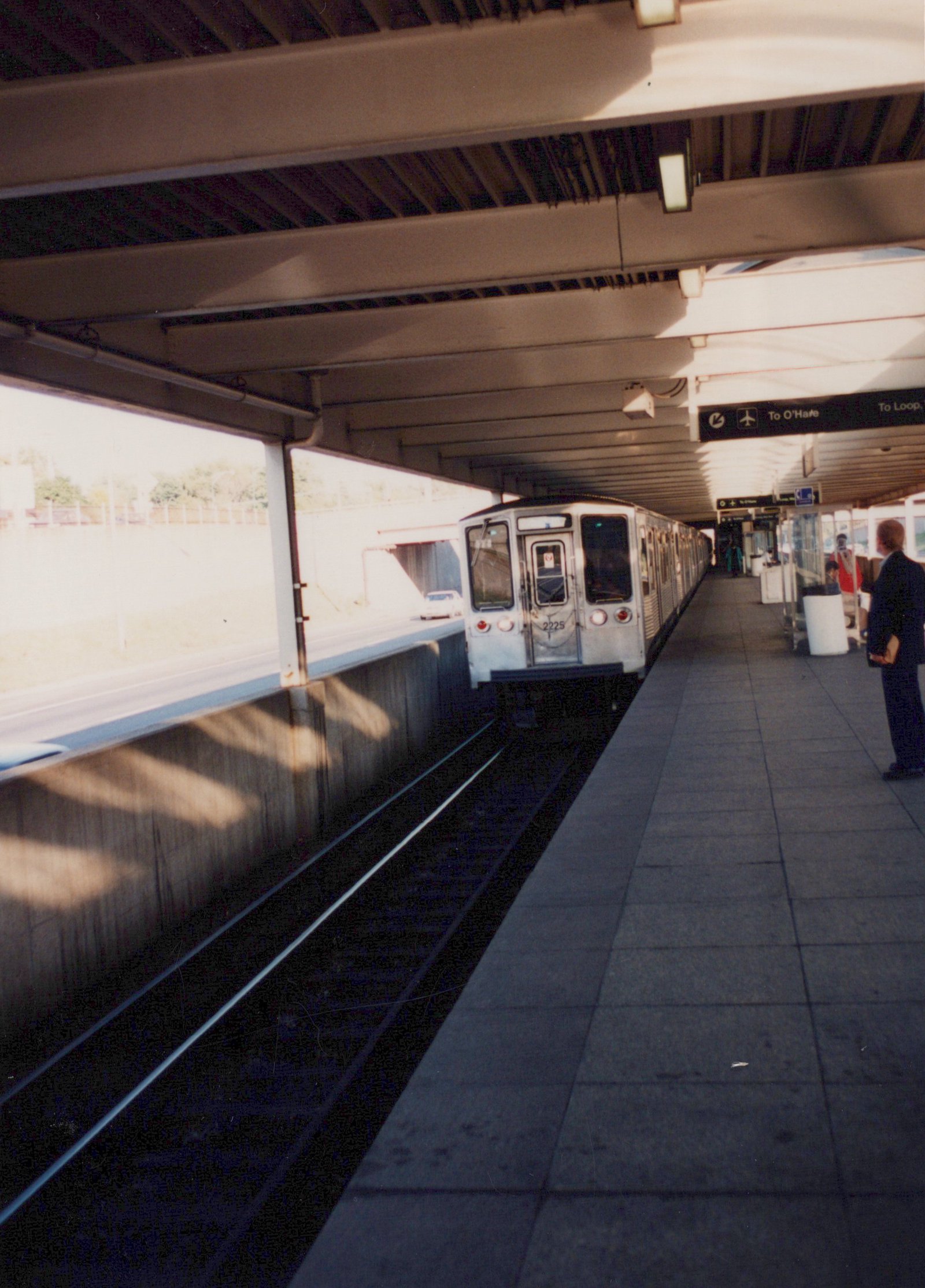 A silver CTA rapid transit train with the number 2225 on its front is stopped at a station platform under a covered structure; signs above the platform indicate directions to O'Hare and the Loop.