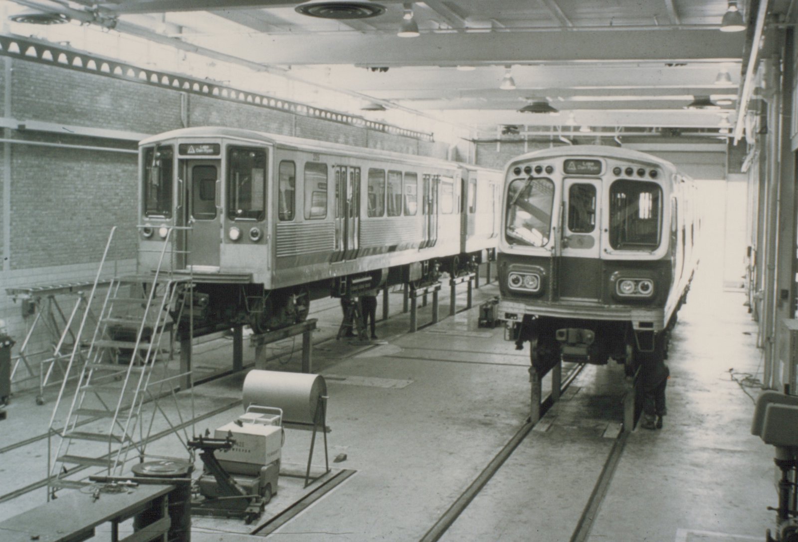 Two CTA rapid transit cars on maintenance tracks inside a large industrial building, with ladders and equipment nearby.