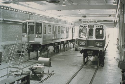 Two CTA rapid transit cars on maintenance tracks inside a large industrial building, with ladders and equipment nearby.