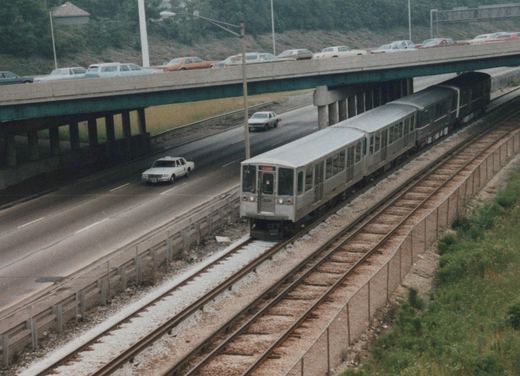 A silver CTA rapid transit train with the number 3222 on its front moves along tracks beside a highway with 1980s-era cars, under an overpass, on a cloudy day in Chicago.