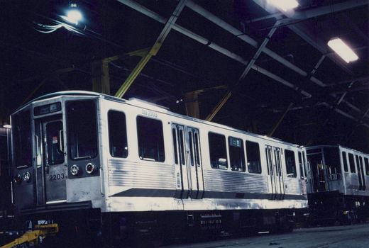 A white and black CTA 2200-series rapid transit car, numbered 2203, is parked indoors in a dimly lit shop with its destination sign reading 'O'Hare Doubles'.