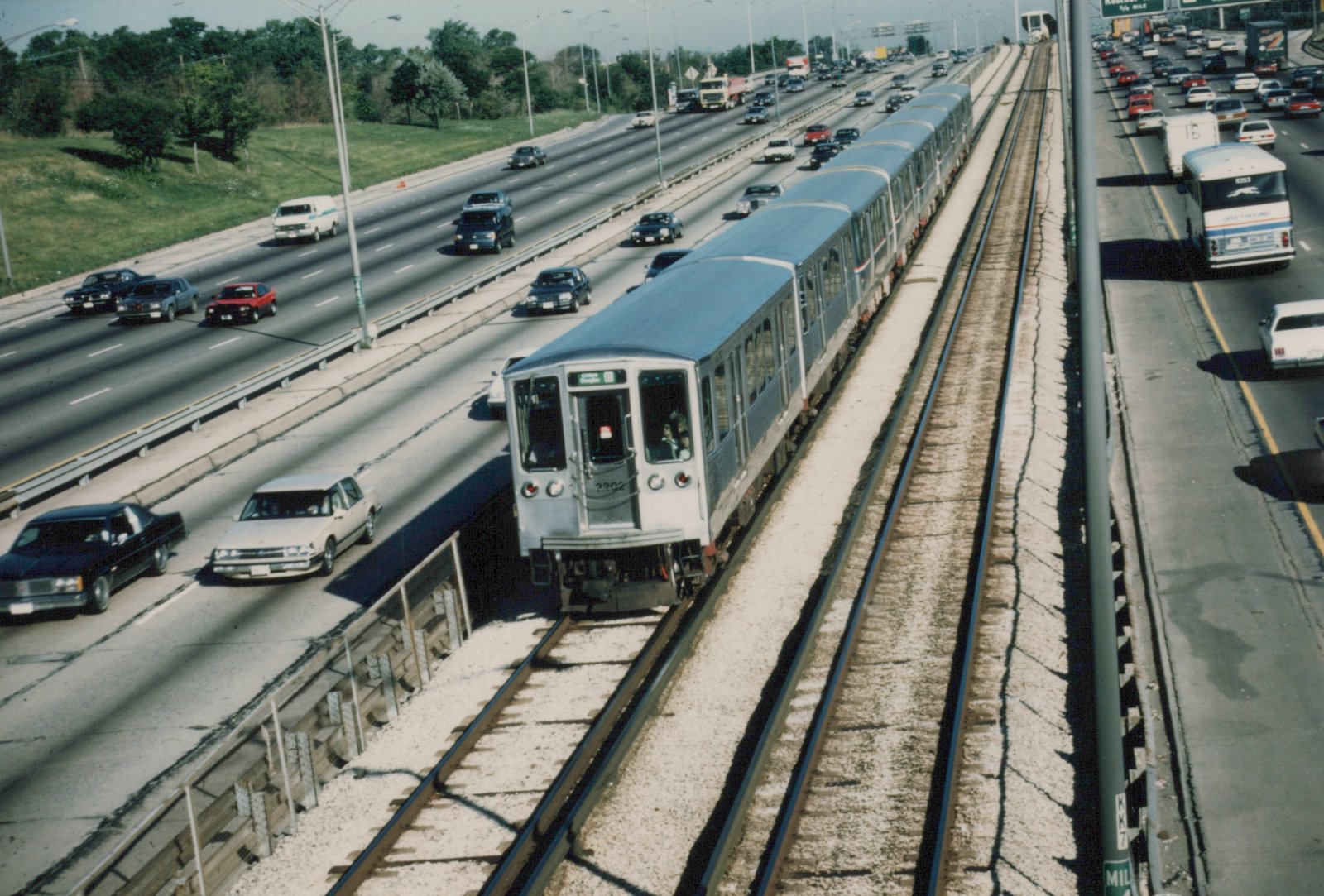 A silver and blue Chicago 'L' train, car number 2202, runs on tracks in the median of a busy expressway, with cars and a Greyhound bus visible on the road.