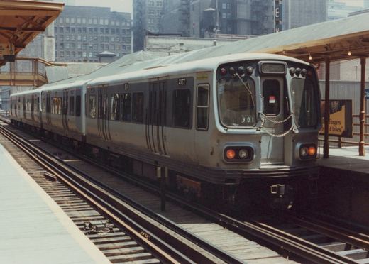 A silver Chicago Transit Authority rapid transit car, number 2177, is stopped at an elevated station platform on a clear day, with city buildings in the background.