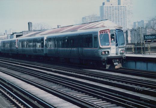 A silver CTA train with red and blue stripes, car number 2081, stopped at an elevated station platform with a sign that reads Addison; the background shows city buildings under an overcast sky.