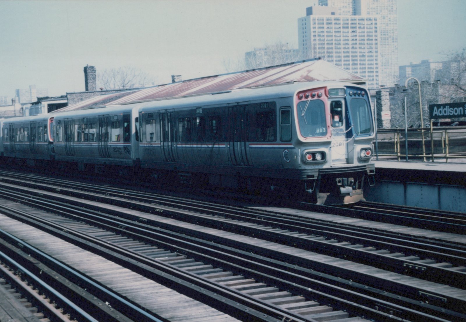 A silver CTA train with red and blue stripes, car number 2081, stopped at an elevated station platform with a sign that reads Addison; the background shows city buildings under an overcast sky.