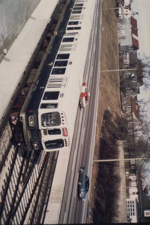 An overhead view of a white and black CTA rapid transit car, number 209, on an elevated track next to a highway with vintage cars, in a winter scene.