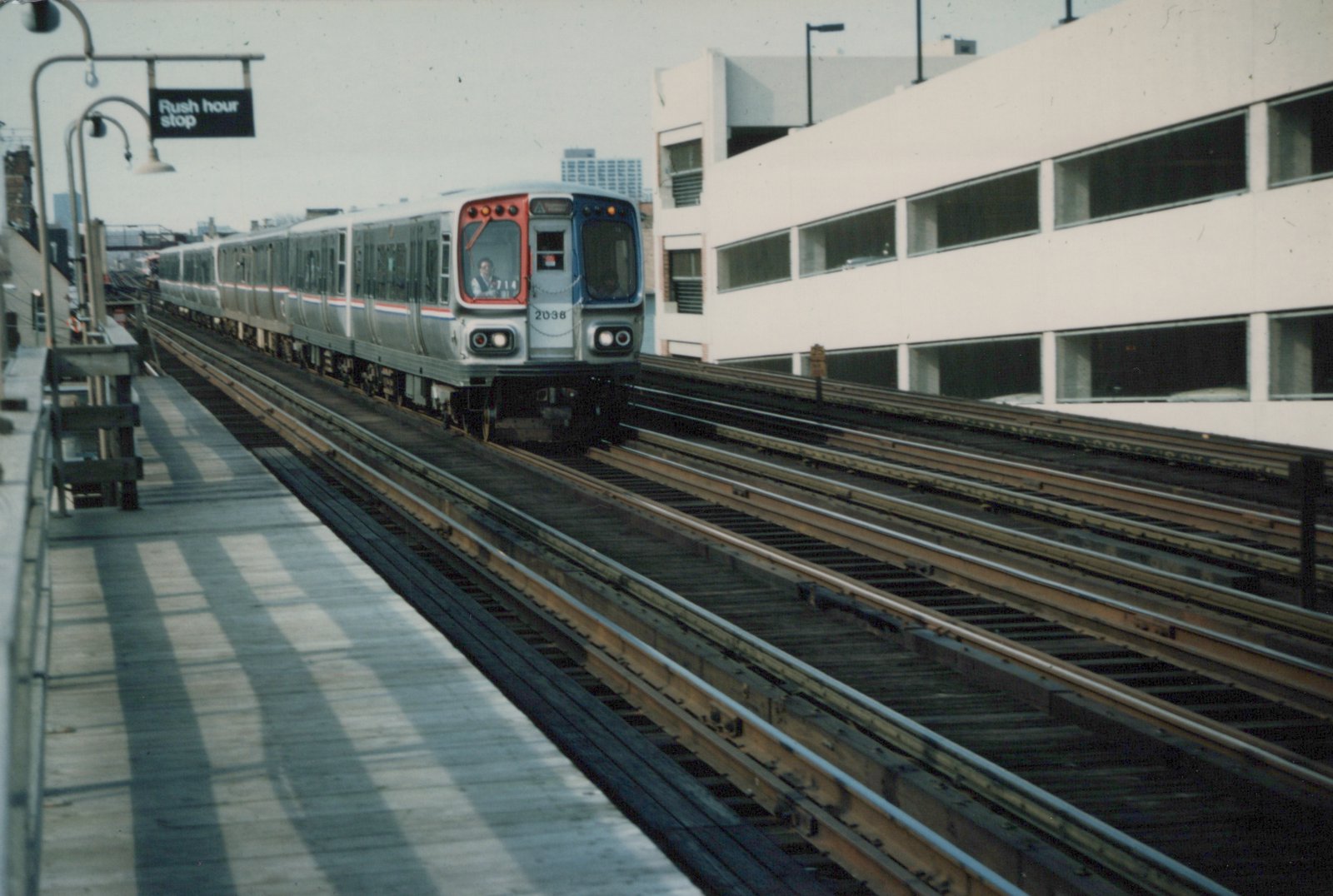 A silver CTA rapid transit train with red and blue stripes, numbered 2038, travels on elevated tracks beside a station platform under a clear sky.