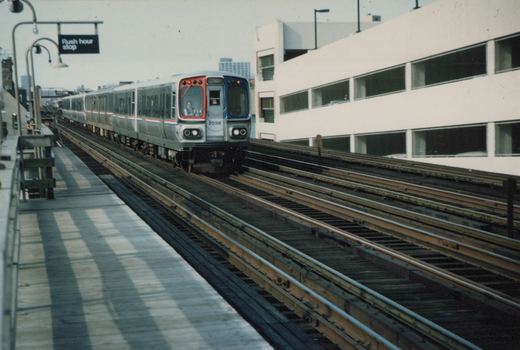 A silver CTA rapid transit train with red and blue stripes, numbered 2038, travels on elevated tracks beside a station platform under a clear sky.