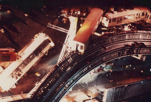 A nighttime overhead shot of a derailed white CTA train on an elevated track, with a fire truck below and emergency lights illuminating the scene.