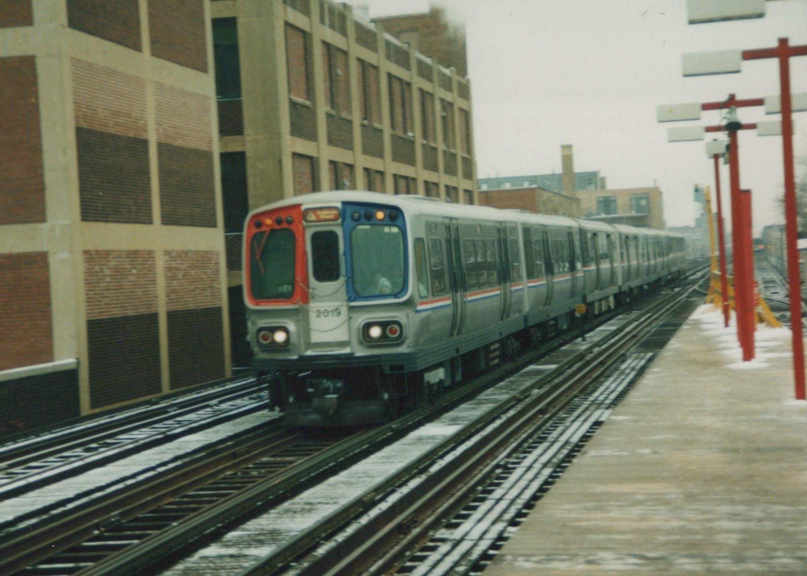 A silver CTA 2000-series subway train, number 2019, with red and blue window trim, is at an elevated station platform with snow on the ground and a large brick building beside the tracks.