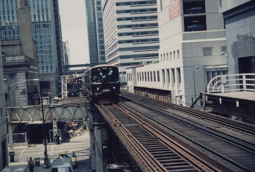 A dark green and maroon CTA 'L' train numbered 185 on an elevated track in a city, with tall buildings and street traffic below on a clear day.