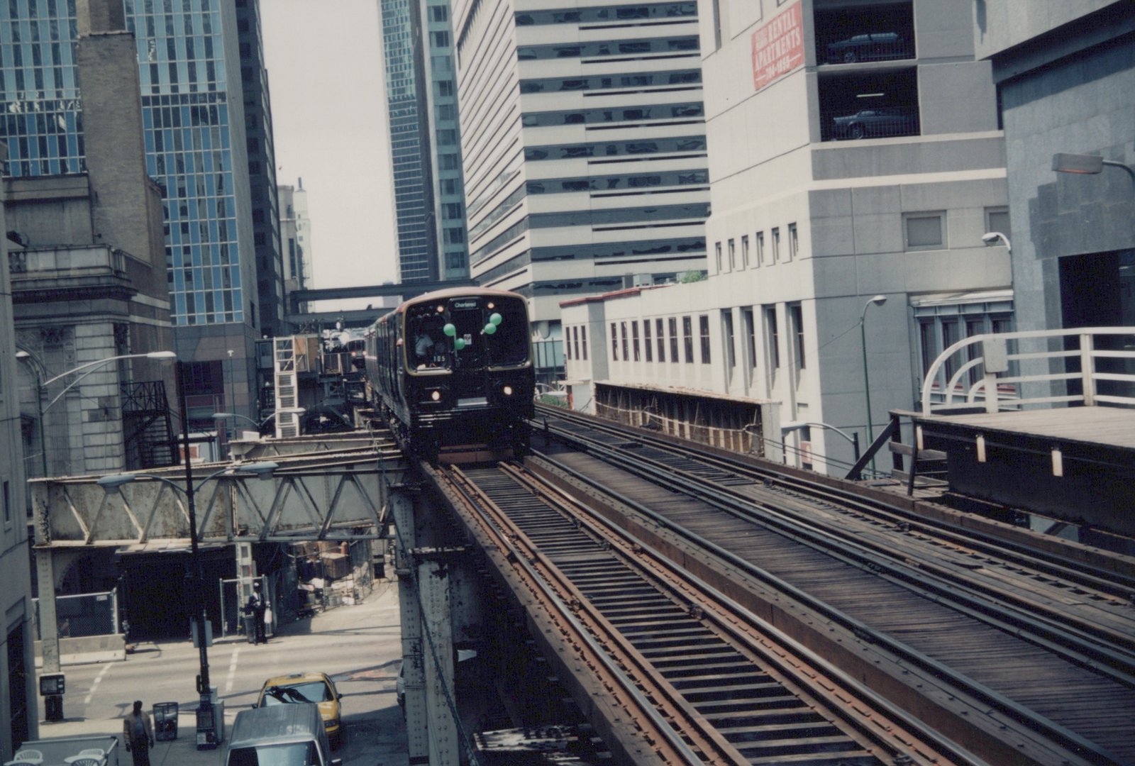 A dark green and maroon CTA 'L' train numbered 185 on an elevated track in a city, with tall buildings and street traffic below on a clear day.
